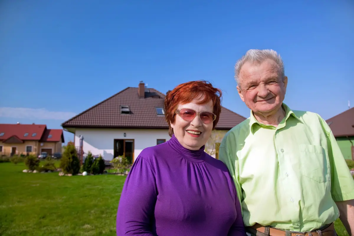 Fotografia przestawia starsze małżeństwo na tle domu jednorodzinnego.